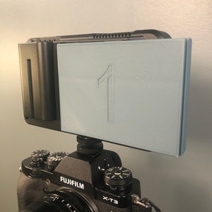 May include: A black Fujifilm X-T3 camera with a lens attached. A gray, rectangular accessory with the number "1" printed on it is attached to the top of the camera.