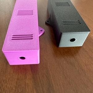 May include: Two rectangular boxes, one purple and one black, with a small hole in the centre of each. Both boxes have a textured surface and multiple small rectangular openings.