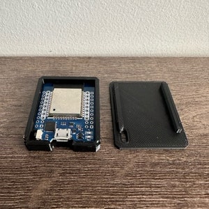 Puede incluir: Una caja electrónica rectangular negra abierta con una placa de circuito azul y una tapa negra separada. La placa de circuito tiene varios componentes, incluido un chip central y múltiples puntos de conexión. El dispositivo está sobre una superficie de madera.