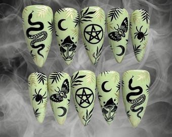May include: Ten green and gold false nails with black designs. The designs include a snake, a crescent moon, a pentagram, a mushroom, a butterfly, and a spider.