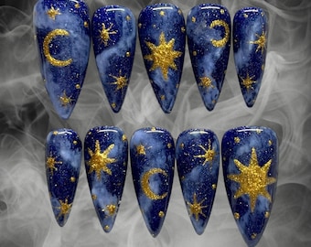 Könnte beinhalten: Set mit zehn blauen und goldenen Nagelspitzen mit einem himmlischen Thema. Jede Spitze hat ein anderes Design mit Sternen, Monden und Sonnen.