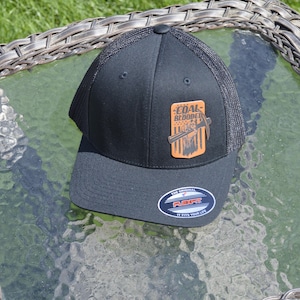 May include: A black baseball cap with a mesh back and a flat brim. The front features a rectangular orange patch with a graphic of a pickaxe and the text "COAL BLOODED" over a striped pattern. A circular sticker on the brim reads "THE ORIGINAL FLEXFIT".