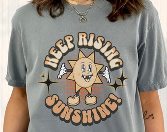 May include: Gray t-shirt with a retro-style sun graphic and the text "Keep Rising Sunshine!"