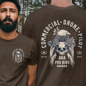 May include: A brown t-shirt with a graphic of a skull with an eagle perched on top. The text "Commercial Drone Pilot" is written in a circle around the graphic. The text "AKA Pro Bird Dodger" is written below the graphic.