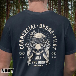 May include: Navy blue t-shirt with a vintage-style graphic of an eagle with a skull, surrounded by roses and a sunburst. The text reads "Commercial Drone Pilot AKA Pro Bird Dodger".