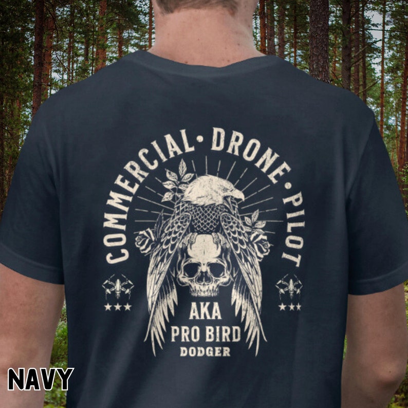 May include: Navy blue t-shirt with a vintage-style graphic of an eagle with a skull, surrounded by roses and a sunburst. The text reads "Commercial Drone Pilot AKA Pro Bird Dodger".