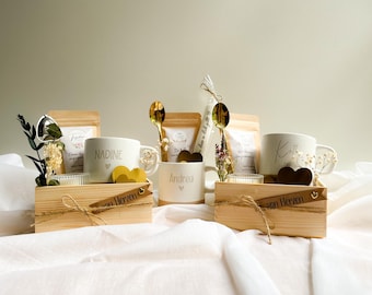 Pode incluir: Conjuntos de presente em caixas de madeira, cada um contendo uma caneca personalizada, uma colher dourada, chá, uma vela e elementos decorativos. As canecas brancas têm nomes como "Nadine", "Andrea" e "Kathie". As caixas têm uma fita que diz "von Herzen".