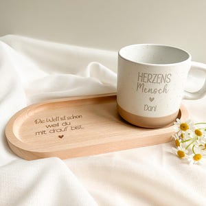 Könnte beinhalten: Eine weiß gesprenkelte Keramiktasse mit den Aufschriften "Herzens Mensch" und "Dani", die auf einem Holztablett steht. Auf dem Tablett steht der eingravierte Text "Die Welt ist schon weil du mit drauf bist." Ein kleiner Strauß weißer Blumen liegt neben dem Tablett.