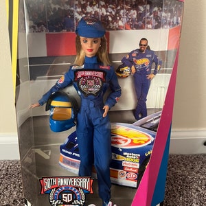 May include: A Barbie doll wearing a blue racing suit with the 50th Anniversary NASCAR logo. The doll is holding a blue helmet and is standing in front of a red, white, and blue race car. The box is labeled "Collector Edition" and "50th Anniversary NASCAR Barbie".