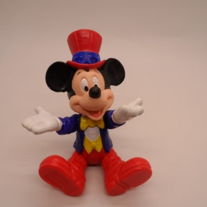 May include: A red, white, and blue Mickey Mouse figurine wearing a red, white, and blue hat. The figurine is smiling and has its arms outstretched.