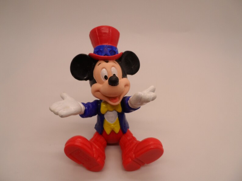 May include: A red, white, and blue Mickey Mouse figurine wearing a red, white, and blue hat. The figurine is smiling and has its arms outstretched.