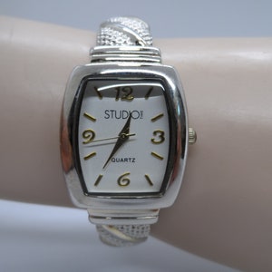 Peut inclure: Montre argentée avec un cadran blanc et des accents dorés. Le cadran de la montre indique "Studio Time" et "Quartz". La montre a un bracelet argenté de style bracelet.