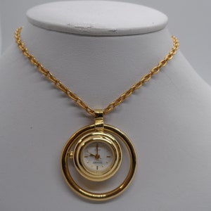 Puede incluir: Un collar de cadena dorada con un colgante dorado que presenta una esfera de reloj redonda con una esfera blanca y agujas negras. La esfera del reloj está rodeada por un anillo dorado más grande.