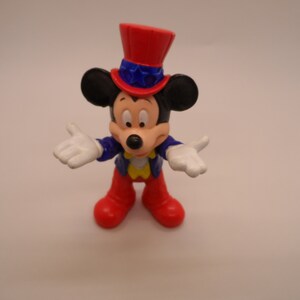 May include: A red, white, and blue Mickey Mouse figurine wearing a red, white, and blue top hat with stars. The figurine is holding out both hands, palms up.