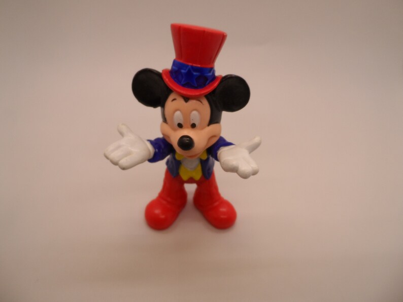 May include: A red, white, and blue Mickey Mouse figurine wearing a red, white, and blue top hat with stars. The figurine is holding out both hands, palms up.