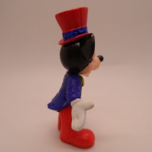 May include: A red, white, and blue plastic figurine of Mickey Mouse wearing a red top hat with blue stars and a blue jacket with white stars. He is wearing red pants and white gloves.