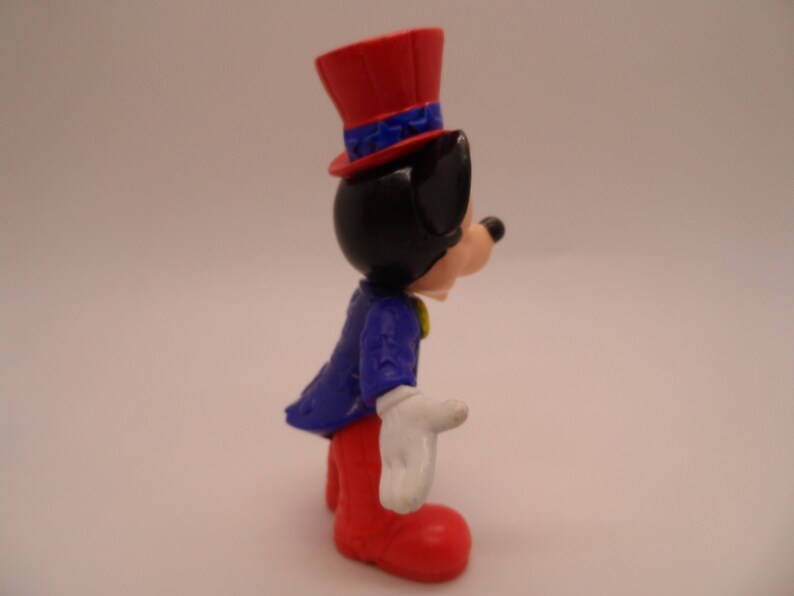 May include: A red, white, and blue plastic figurine of Mickey Mouse wearing a red top hat with blue stars and a blue jacket with white stars. He is wearing red pants and white gloves.