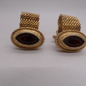 May include: A pair of gold cufflinks with a red oval-shaped stone in the center. The cufflinks have a chain-link design.