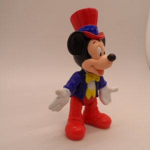 May include: A red, white, and blue plastic figurine of Mickey Mouse wearing a top hat and a blue jacket with a yellow bow tie.