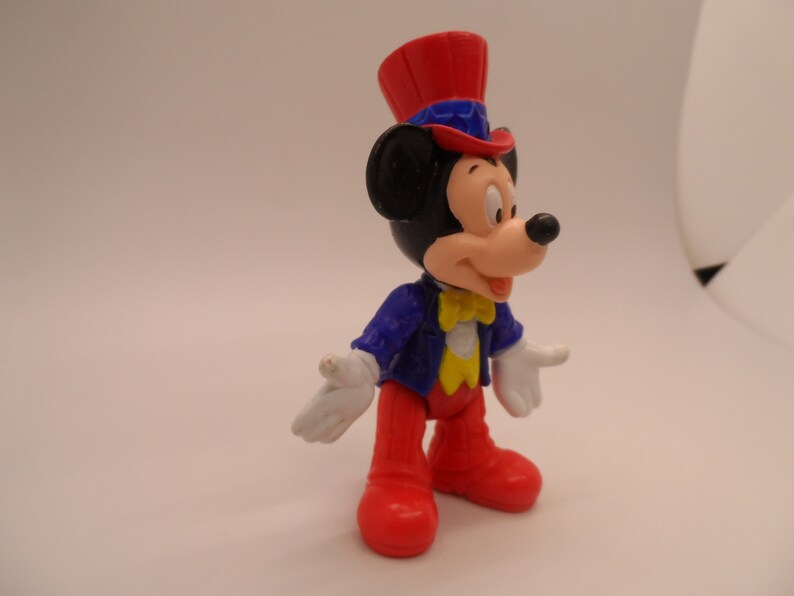 May include: A red, white, and blue plastic figurine of Mickey Mouse wearing a top hat and a blue jacket with a yellow bow tie.