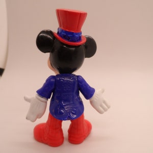 May include: A black and white Mickey Mouse figurine wearing a red, white, and blue top hat and a blue jacket. The figurine is standing on a white background.