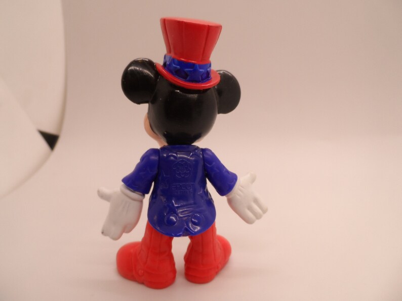 May include: A black and white Mickey Mouse figurine wearing a red, white, and blue top hat and a blue jacket. The figurine is standing on a white background.