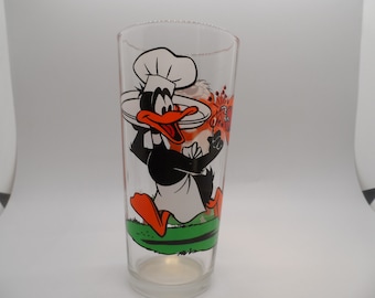 May include: A glass drinking glass featuring a cartoon image of Daffy Duck dressed as a chef. He is holding a plate and a spatula. The glass is clear and has a green and orange design.