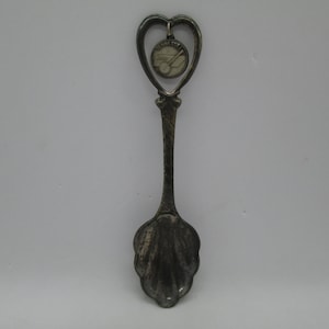 May include: Silver souvenir spoon with a heart-shaped handle and a charm featuring a small image of a train. The charm is attached to the handle with a small ring.