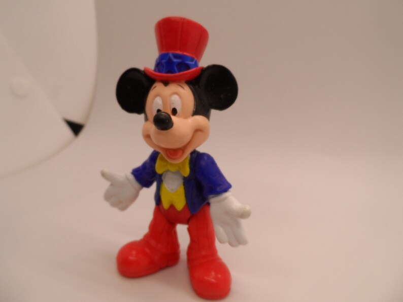 May include: A red, white, and blue Mickey Mouse figurine wearing a red, white, and blue Uncle Sam hat. The figurine is made of plastic and has a yellow bow tie.