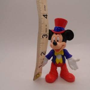 May include: A plastic Mickey Mouse figurine wearing a red, white, and blue top hat with a star on it. He is wearing a blue jacket with a yellow bow tie and red pants. He is about 3 inches tall.