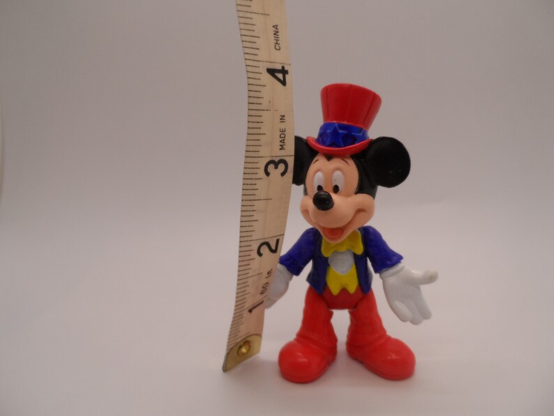 May include: A plastic Mickey Mouse figurine wearing a red, white, and blue top hat with a star on it. He is wearing a blue jacket with a yellow bow tie and red pants. He is about 3 inches tall.