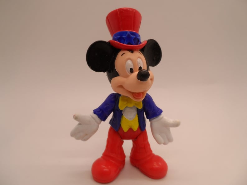May include: A red, white, and blue plastic Mickey Mouse figurine wearing a red, white, and blue top hat. The figurine is wearing a blue jacket with yellow accents and red pants.
