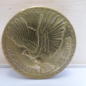 May include: A gold-colored coin with the words "REPUBLICA DE CHILE" around the edge. The coin features a stylized image of a condor with its wings spread.