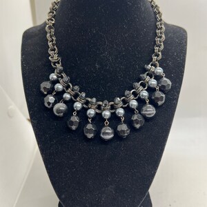 May include: A black and silver necklace with a chain and large black beads. The necklace is displayed on a black mannequin bust.