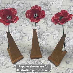 May include: Three red poppy sculptures with black centres and green stems are displayed on wooden stands. The stands are triangular and made of light brown wood. The background is a world map. Text at the bottom reads: Poppies shown are for illustration purposes only.