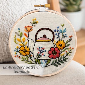 Può includere: Ricamo di un motivo floreale a forma di teiera su tessuto color crema, teso all'interno di un telaio di legno. La teiera è bianca con manico e beccuccio marroni, circondata da fiori colorati gialli, arancioni e rossi. Testo: "Embroidery pattern template".