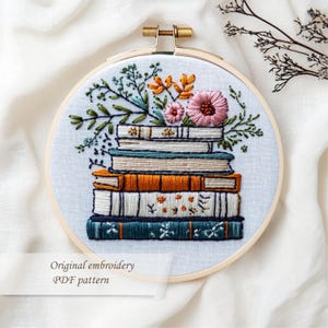 May include: Embroidered hoop art featuring a stack of books with colorful spines, topped with pink and orange flowers and green foliage. The text "Original embroidery PDF pattern" is below the design. The embroidery is set in a wooden hoop.