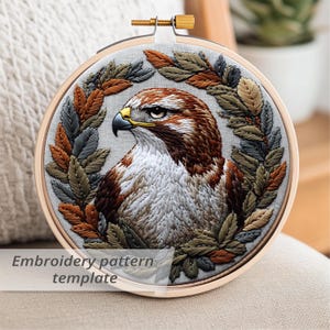May include: Round embroidery hoop featuring a detailed embroidered hawk with brown, white, and gray feathers, surrounded by a wreath of green, brown, and gray leaves. The text "Embroidery pattern template" is visible at the bottom.