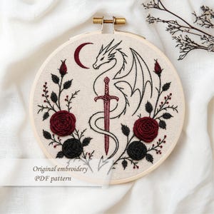 May include: Embroidered hoop art featuring a dragon, sword, and crescent moon design. The artwork includes red and black roses and the text "Original embroidery PDF pattern". The embroidery is set in a wooden hoop.