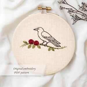 May include: An embroidery hoop featuring a black crow perched on a branch with red roses and green leaves. The text "Original embroidery PDF pattern" is displayed on a white label. The hoop is held by a gold-colored metal clasp.