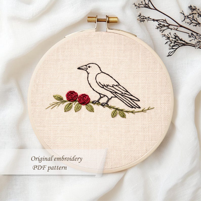 May include: An embroidery hoop featuring a black crow perched on a branch with red roses and green leaves. The text "Original embroidery PDF pattern" is displayed on a white label. The hoop is held by a gold-colored metal clasp.