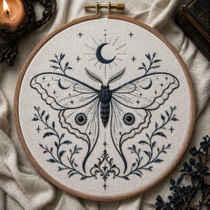 Puede incluir: Bordado en un aro de madera con un diseño detallado de polilla, una luna creciente y estrellas. La polilla es azul oscuro, con intrincados patrones de alas y elementos florales. El fondo es de tela beige claro.