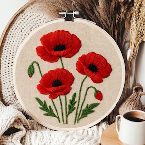 May include: A round embroidery hoop featuring a detailed floral design. Three vibrant red poppies with black centers are embroidered on a beige fabric. Green leaves and stems complete the botanical artwork, creating a decorative piece.