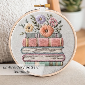 May include: A round embroidery hoop featuring a stack of embroidered books with colorful floral accents. The design includes a variety of flowers in shades of peach, lavender, and yellow. The text "Embroidery pattern template" is visible at the bottom.