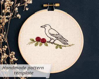 May include: An embroidery hoop with a crow design. The crow is perched on a branch with red roses and green leaves. The text "Handmade pattern template" is displayed on a white banner.