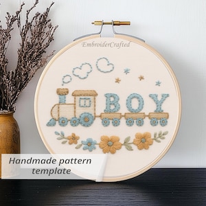 Op de afbeelding: Geborduurde hoepelkunst met een trein en het woord "BOY" in blauwe draad. De trein is bruin en blauw, met geborduurde wolken en bloemen. De tekst "Handmade pattern template" is zichtbaar.