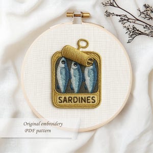 May include: Embroidered artwork of a sardine can with three blue fish. The embroidery is set in a wooden hoop. The word "SARDINES" is embroidered on the can. The text "Original embroidery PDF pattern" is below the hoop.