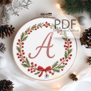 Puede incluir: Arte de aro bordado con la letra "A" en cursiva, rodeada por una corona de hojas verdes y bayas rojas. Un lazo rojo en la parte inferior. El texto "PDF EmbroiderCrafted" es visible.