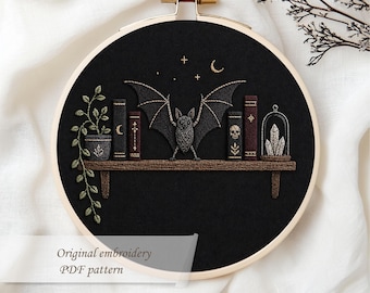 May include: Embroidered hoop art featuring a black background with a wooden shelf. The shelf holds a potted plant, books, a bat, and a glass cloche with crystals. The design includes a crescent moon and stars. The text "Original embroidery PDF pattern" is at the bottom.