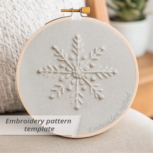 May include: A light grey embroidery hoop featuring a snowflake design. The text "Embroidery pattern template" is visible. The wooden hoop is secured with a brass screw. The design is approximately 15 cm in diameter.